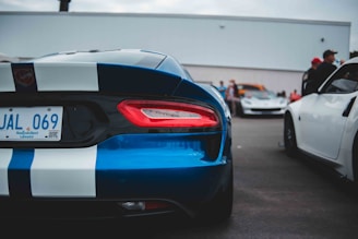 A parking lot scene with a focus on a blue sports car with white racing stripes. The rear end of the car is prominently displayed, showcasing its taillights and license plate. Other vehicles and people in the background suggest a car gathering or event.