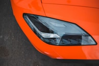 Close-up of a sleek car headlight set against a vibrant orange body. The headlight features modern design with sharp lines and a reflective surface, situated on a dark asphalt background.