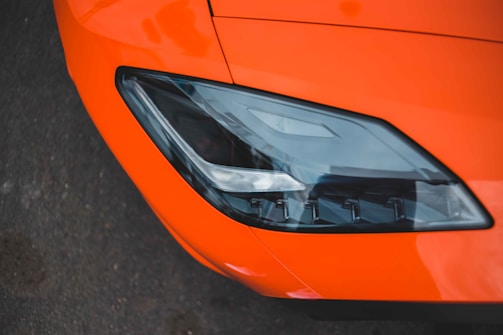 Close-up of a sleek car headlight set against a vibrant orange body. The headlight features modern design with sharp lines and a reflective surface, situated on a dark asphalt background.