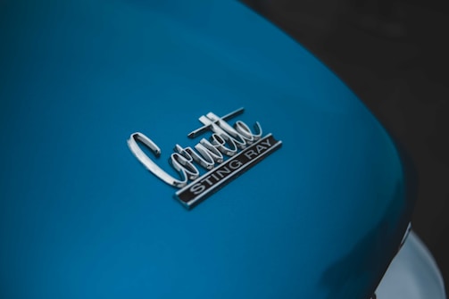 A close-up view of a car's surface displaying the badge with the words 'Corvette Sting Ray' in chrome. The surface is painted in a blue color, with a glossy finish.