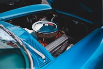 An open hood of a vintage car reveals a clean, well-maintained engine. The engine has a shiny, circular air filter cover with a visible brand name embossed on the valve cover and hints of red engine components beneath. The car's body is painted in a vibrant blue color, adding a striking contrast.