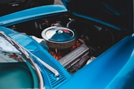 An open hood of a vintage car reveals a clean, well-maintained engine. The engine has a shiny, circular air filter cover with a visible brand name embossed on the valve cover and hints of red engine components beneath. The car's body is painted in a vibrant blue color, adding a striking contrast.