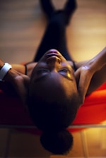 A person is lying back on a red chair with eyes closed, hands resting on the back of their head. The room is softly lit, creating a warm and relaxed atmosphere. The angle of view is from above, emphasizing the sense of tranquility.