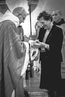 A person in clerical attire hands an object to a smiling woman. Another person stands in the background, wearing ceremonial clothing. The setting appears to be a church or religious space.