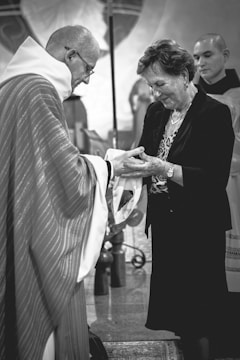A person in clerical attire hands an object to a smiling woman. Another person stands in the background, wearing ceremonial clothing. The setting appears to be a church or religious space.