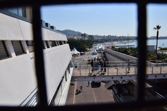A waterfront walkway with people walking and gathering under clear blue skies. Several white tents and palm trees are visible along the path, leading towards a beach area in the distance. The scene is framed by a metal railing, suggesting a view from a higher vantage point.