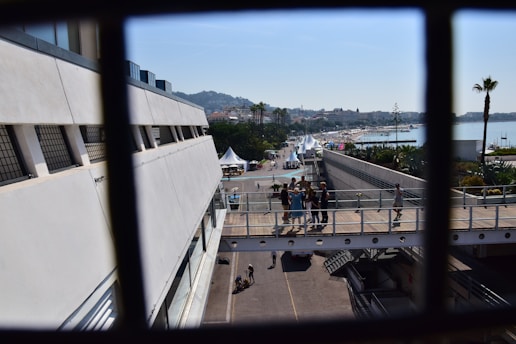 A waterfront walkway with people walking and gathering under clear blue skies. Several white tents and palm trees are visible along the path, leading towards a beach area in the distance. The scene is framed by a metal railing, suggesting a view from a higher vantage point.