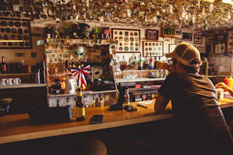 A dimly lit bar with a variety of objects and memorabilia on the walls and ceiling, including currency notes hanging from above. There is a man wearing a cap sitting at the bar, drinking from a bottle. On the bar counter, several bottles, a mug, and other items are visible. The background displays a large collection of framed pictures and mirrors.