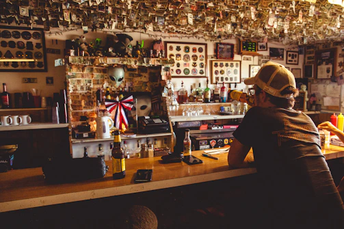 A dimly lit bar with a variety of objects and memorabilia on the walls and ceiling, including currency notes hanging from above. There is a man wearing a cap sitting at the bar, drinking from a bottle. On the bar counter, several bottles, a mug, and other items are visible. The background displays a large collection of framed pictures and mirrors.