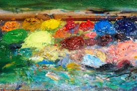 A vibrant array of paint colors is spread across a mixing palette. Blobs of green, yellow, red, blue, orange, and pink are mixed together in an artistic chaos. The rich textures show the thick application of paint, implying a work in progress. The background consists of a wooden border, suggesting the palette is well-used.