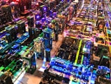 A complex array of computer motherboards and circuit boards is organized in a sprawling grid layout. Bright, multicolored LED lights illuminate the scene, casting vibrant reflections and creating an intricate, high-tech pattern. Wires and electronic components are interwoven, contributing to an aesthetic that resembles a miniature cityscape.