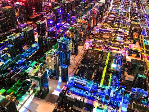 A complex array of computer motherboards and circuit boards is organized in a sprawling grid layout. Bright, multicolored LED lights illuminate the scene, casting vibrant reflections and creating an intricate, high-tech pattern. Wires and electronic components are interwoven, contributing to an aesthetic that resembles a miniature cityscape.
