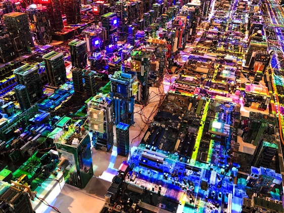 A complex array of computer motherboards and circuit boards is organized in a sprawling grid layout. Bright, multicolored LED lights illuminate the scene, casting vibrant reflections and creating an intricate, high-tech pattern. Wires and electronic components are interwoven, contributing to an aesthetic that resembles a miniature cityscape.