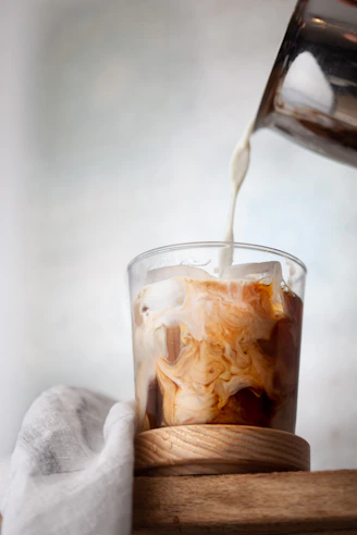Milk pouring into a glass of iced coffee on a wooden coaster