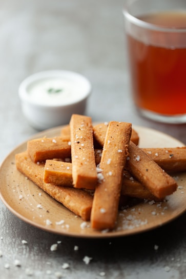 Rectangular pieces of fried or baked snacks are stacked on a ceramic plate, sprinkled with coarse salt. A small bowl of creamy white dipping sauce with herbs sits in the background, alongside a glass of amber-colored tea.