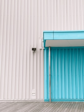 A corrugated metal wall combining white and turquoise panels, featuring a small overhang above the turquoise section. On the left, a surveillance camera is mounted. The ground consists of wooden planks laid horizontally.