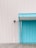 A corrugated metal wall combining white and turquoise panels, featuring a small overhang above the turquoise section. On the left, a surveillance camera is mounted. The ground consists of wooden planks laid horizontally.