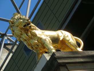 A golden lion statue stands prominently against a backdrop of a modern architectural structure with metal beams and green panels. The lion's mane is detailed and the statue is positioned on a stone pedestal.