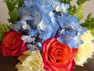 A beautiful arrangement of flowers featuring vivid blue hydrangeas, vibrant red and yellow roses, and lush green foliage.