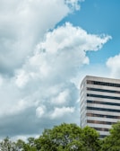A tall, modern office building with a striped glass exterior stands against a background of lush green trees. Large, fluffy clouds fill the sky, which is a bright shade of blue.