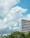 A tall, modern office building with a striped glass exterior stands against a background of lush green trees. Large, fluffy clouds fill the sky, which is a bright shade of blue.