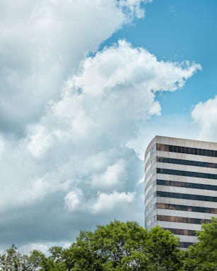A tall, modern office building with a striped glass exterior stands against a background of lush green trees. Large, fluffy clouds fill the sky, which is a bright shade of blue.