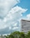 A tall, modern office building with a striped glass exterior stands against a background of lush green trees. Large, fluffy clouds fill the sky, which is a bright shade of blue.