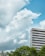 A tall, modern office building with a striped glass exterior stands against a background of lush green trees. Large, fluffy clouds fill the sky, which is a bright shade of blue.