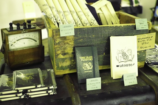 A collection of vintage and themed items are displayed on a table. There is a wooden crate holding rolled-up sheets with decorative designs. Next to it is a small wooden device with a dial, possibly an antique voltmeter. Several books or booklets titled 'TIME TRAVEL PASSPORT' are arranged in stacks, and some are individually displayed. There are various small price tags and product descriptions in Japanese displayed on and around the items.