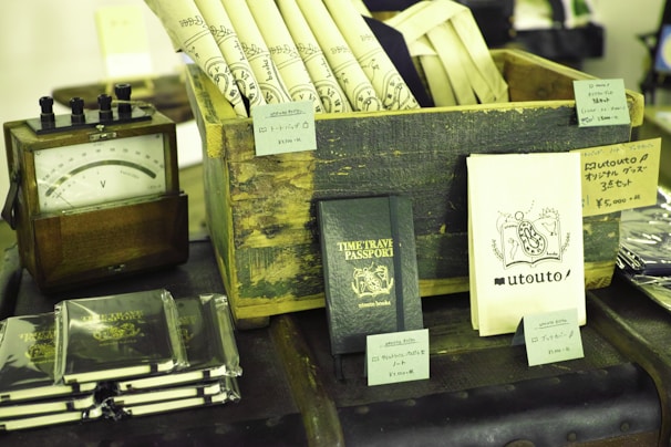 A collection of vintage and themed items are displayed on a table. There is a wooden crate holding rolled-up sheets with decorative designs. Next to it is a small wooden device with a dial, possibly an antique voltmeter. Several books or booklets titled 'TIME TRAVEL PASSPORT' are arranged in stacks, and some are individually displayed. There are various small price tags and product descriptions in Japanese displayed on and around the items.