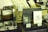 A collection of vintage and themed items are displayed on a table. There is a wooden crate holding rolled-up sheets with decorative designs. Next to it is a small wooden device with a dial, possibly an antique voltmeter. Several books or booklets titled 'TIME TRAVEL PASSPORT' are arranged in stacks, and some are individually displayed. There are various small price tags and product descriptions in Japanese displayed on and around the items.