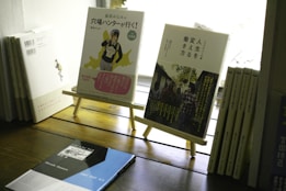 A selection of books is displayed on a wooden surface. Two books stand on wooden easels, featuring colorful and artistic covers with Japanese text. Additional books are stacked and lined up, creating a cozy and literary ambiance.