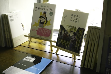A selection of books is displayed on a wooden surface. Two books stand on wooden easels, featuring colorful and artistic covers with Japanese text. Additional books are stacked and lined up, creating a cozy and literary ambiance.