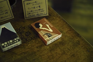 A wooden surface displays two retro-styled matchboxes with vintage Japanese designs. One matchbox features an illustration of a woman in profile, elegantly dressed and holding a cigarette. The other box displays an image of Mount Fuji with Japanese text along the bottom. Handwritten signs in Japanese, likely offering details or prices, are partially visible in the background.
