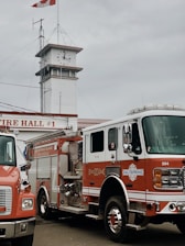 A fire station featuring a clock tower with a winding staircase and a prominent Canadian flag on top. The building is marked as 'Fire Hall #1'. Two fire trucks are parked in front, one bearing the label 'Salt Spring Island Fire Rescue'. The scene is set against a cloudy sky.