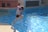 A child wearing a life vest jumps into a swimming pool with sparkling blue water. Sunlight reflects off the surface, creating bright spots. The background shows part of the pool area with warm toned walls and windows.