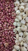 A display of red onions on the left side, contrasting with a pile of white garlic heads on the right. The onions are small, round, and tightly packed together, showcasing their reddish-purple skin. The garlic heads are slightly larger, with their papery white skin and some stems attached.