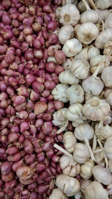 A display of red onions on the left side, contrasting with a pile of white garlic heads on the right. The onions are small, round, and tightly packed together, showcasing their reddish-purple skin. The garlic heads are slightly larger, with their papery white skin and some stems attached.