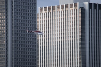 A helicopter with red and white colors is flying in front of tall urban skyscrapers. The buildings have a grid-like pattern with numerous windows and vertical lines, suggesting a modern architectural style.