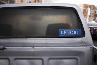 A vehicle with a slightly rusted rear and a sticker on the back window that reads 'Vote for Obi-Wan Kenobi Our Only Hope' in blue and white. The background includes blurred urban elements.