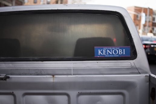A vehicle with a slightly rusted rear and a sticker on the back window that reads 'Vote for Obi-Wan Kenobi Our Only Hope' in blue and white. The background includes blurred urban elements.