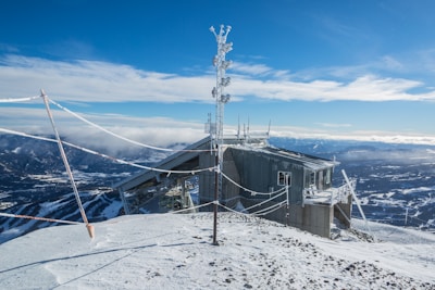A snow-covered mountainous landscape features a small, rugged building with metal siding and roof. Several antennas and communication equipment are mounted on the building. The sky is clear and blue with some clouds hovering over distant peaks. Snow and ice are visible on the ropes and structure, indicating a cold climate.