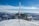 A snow-covered mountainous landscape features a small, rugged building with metal siding and roof. Several antennas and communication equipment are mounted on the building. The sky is clear and blue with some clouds hovering over distant peaks. Snow and ice are visible on the ropes and structure, indicating a cold climate.