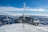 A snow-covered mountainous landscape features a small, rugged building with metal siding and roof. Several antennas and communication equipment are mounted on the building. The sky is clear and blue with some clouds hovering over distant peaks. Snow and ice are visible on the ropes and structure, indicating a cold climate.
