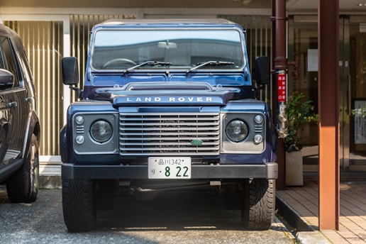 A blue Land Rover vehicle is parked in a driveway or parking space, surrounded by other cars and urban elements. The front view emphasizes its distinct grille and headlights. A plant and metal post are visible on the right side.