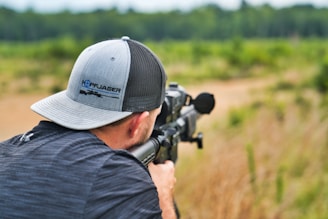 A person is aiming a rifle outdoors, focusing on a target. The setting is a natural environment with green foliage in the background. The individual is wearing a two-tone cap and a black shirt.