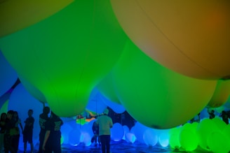 A group of large, colorful inflatable spheres, predominantly in shades of green, yellow, and blue, dominates the space. People stand and walk around these glowing spheres, creating a vibrant, immersive environment. The lighting casts a surreal and playful atmosphere.