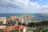 A panoramic view of a coastal city with a mix of high-rise buildings and greenery. In the foreground is a circular bullring, surrounded by apartment buildings and vibrant red roofs. The sea is visible in the background, with a large harbor featuring cranes and cargo ships on the right.