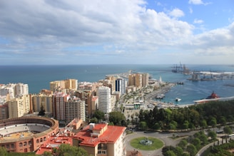 A panoramic view of a coastal city with a mix of high-rise buildings and greenery. In the foreground is a circular bullring, surrounded by apartment buildings and vibrant red roofs. The sea is visible in the background, with a large harbor featuring cranes and cargo ships on the right.