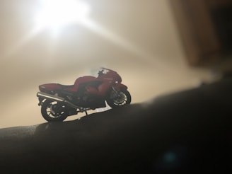 A detailed model of a motorcycle is positioned on a surface in a dimly lit environment. The bike, with its red and black coloration, stands out against the blurred and softly illuminated background. The lighting creates a silhouette effect, enhancing the contours and details of the motorcycle.